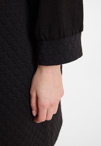 Schwarzes, strukturiertes Kleid mit einem sechseckigen Muster und einem sanften, gerafften Ärmelbündchen. Nahaufnahme einer Hand, die an dem Stoff ruht.