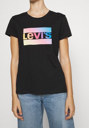 Person iført en sort T-shirt med et farverigt gradient Levi's-logo og lyseblå denimjeans, stående mod en ensfarvet baggrund.