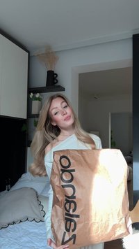 Młoda kobieta z długimi blond włosami trzymająca dużą brązową torbę na zakupy z czarnym napisem, na tle nowoczesnej sypialni.