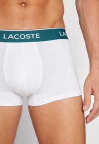 Bijeli pamučni bokserice s tirkiznim elastičnim pojasom na kojem se nalazi logo "LACOSTE". Prilagođen dizajn, mekana tekstura, čisti šavovi.