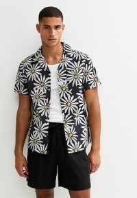 Chemise noire à manches courtes avec un imprimé de marguerites blanches, centres jaunes, et col noté ; associée à un short noir. Matériau en coton, design décontracté.