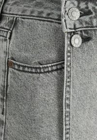 I jeans in denim grigio presentano un primo piano della zona della tasca, con una trama texturizzata e cuciture a contrasto. Sono visibili due bottoni argentati.