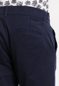 Puuvillaiset tummansiniset chinot, joissa on sileä pinta, takatasku ja vyölenkit, nähtynä sivukulmasta.