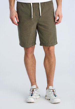 Mann trägt olivgrüne Cord-Shorts mit weißem Kordelzug und weißen Sportschuhen mit schwarzen und roten Details, steht vor einfachem Hintergrund.