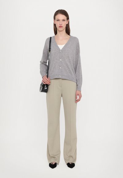 Pelēks adīts kardigāns, ar V veida kakla izgriezumu, ar pogām priekšā, valkāts virs baltas blūzes. Beidz kreisās puses bikses, melnas asās kurpes, minimāla melna soma. Balta fona.