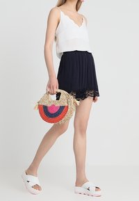 Bílý bezrukávový top s vlnitými okraji, námořnicky modré šortky s krajkovým lemem, barevná tkaná kruhová taška a bílé platformové sandály.