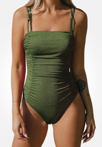 Olivgrüner Einteiler-Badeanzug mit gerafftem Material, schmalen Trägern, quadratischem Ausschnitt und moderatem Beinausschnitt. Dezente Schimmerakzente zieren den Stoff.