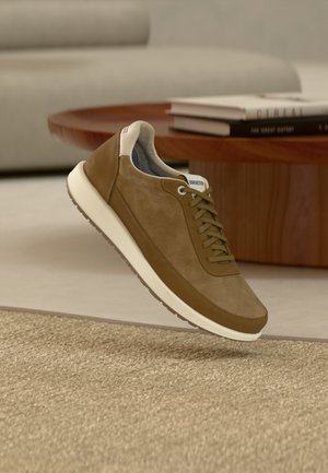 Rjava semiš in usnjena superga z belim podplatom, vezalkami in oznako "Birkenstock", lebdi nad bež preprogo v dnevni sobi.