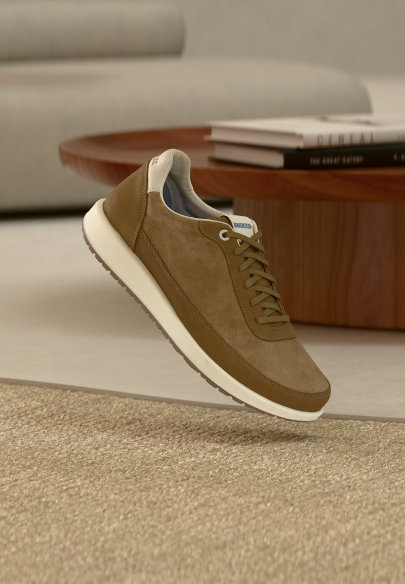 Καφέ σουέντ και δερμάτινο αθλητικό παπούτσι με λευκή σόλα, κορδόνια και ετικέτα "Birkenstock", αιωρούμενο πάνω από μπεζ χαλί σε σαλόνι.