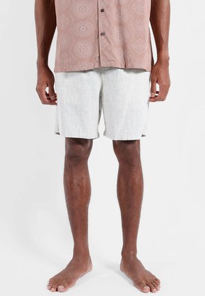 Hombre con pantalones cortos gris claro hasta la rodilla y una camisa rosa con botones y estampado, de pie descalzo sobre un fondo liso.