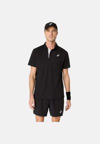 Uomo che indossa un cappellino sportivo nero, una polo nera, pantaloni corti neri e un braccialetto nero, in piedi contro uno sfondo chiaro e uniforme.