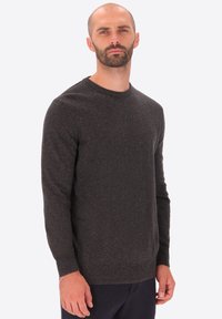 Uomo adulto con barba corta, che indossa un maglione a maniche lunghe grigio scuro e pantaloni scuri, in piedi su uno sfondo chiaro.