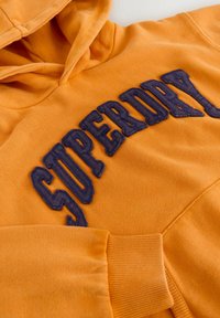 Sudadera naranja con un bolsillo frontal tipo canguro y letras texturizadas en azul marino que dicen "SUPERDRY" en el pecho.