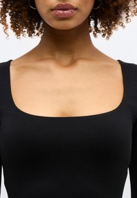 Γυναίκα με κατσαρά μαλλιά που φοράει μαύρο τοπ με φαρδιά τετράγωνη λαιμόκοψη, που δείχνει τα κλειδωματά της και τα χείλη της, σε λευκό φόντο.