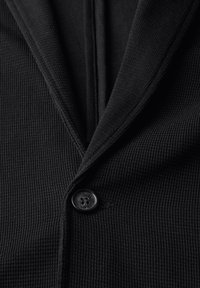 Svart ribbad kofta med en notched krage och en enda knappstängning. Tyget har en strukturerad yta.