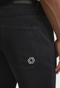 Gros plan sur la poche d'un pantalon noir avec une broderie de logo hexagonal blanc et une partie de la main d'une personne reposant à côté de la jambe.