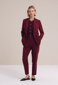 Žena stojící s rukama v kapsách, oblečená ve vínovém obleku, zapnutém kardiganu, perlových náušnicích a černých špičatých balerínách na béžovém pozadí.