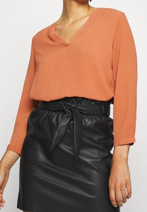 Kobieta ubrana w rdzawobrązową bluzkę z długim rękawem i dekoltem w serek, wpuszczoną w wysokowatową czarną spódnicę z eko skóry z przewiązanym paskiem.