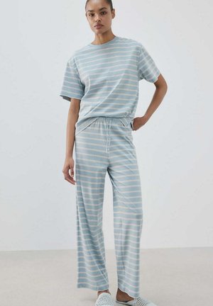 Femme debout, main sur la hanche, portant un t-shirt assorti rayé horizontal bleu clair et blanc et un pantalon à jambes larges avec des chaussures blanches sans lacets.