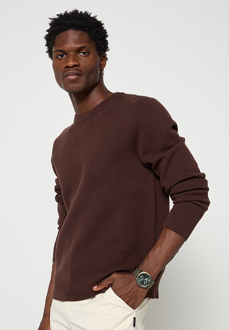 Hombre con un suéter largo marrón y pantalones beige, con la mano en el bolsillo, mostrando un reloj de pulsera, de pie contra un fondo claro y liso.