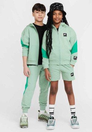 Mintgroene sportkleding bestaat uit een jack met rits en contrasterende mouwen, samen met bijpassende shorts. Accessoires omvatten een gestreepte zwarte bucket hat en sneakers.