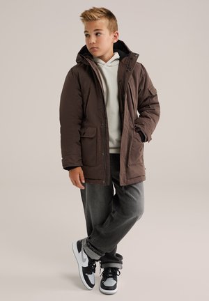 Junge trägt eine braune Winterjacke mit Kapuze, ein beiges Hoodie, graue Jeans und schwarz-weiße Sneaker und steht mit einer Hand in der Tasche.