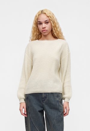 Ung kvinde med langt krøllet blondt hår iført en cremefarvet striktrøje og mørke, løstsiddende jeans, stående mod en ensartet lys baggrund.