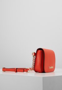 Sac bandoulière en cuir rouge avec un accent de chaîne dorée, forme rectangulaire, rabat pliant et une sangle ajustable. Texture lisse avec des bordures noires.