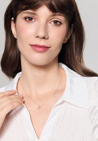 Złoty naszyjnik z sercowym wisiorkiem i małym akcentem kamienia, noszony na białej strukturalnej koszuli. Prosty i elegancki design.