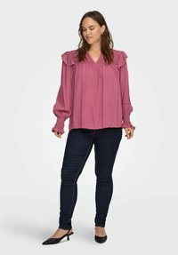 Kobieta nosząca ciemne dopasowane dżinsy, czarne buty na niskim obcasie ze zapięciem na pięcie oraz luźną, różową bluzkę z długim rękawem i falbanami.