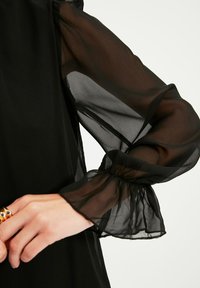 Schwarze Bluse mit transparenten schwarzen Puffärmeln und gerafften Manschetten. Glatter Stoff mit leichter Textur, der das feste Oberteil ergänzt.