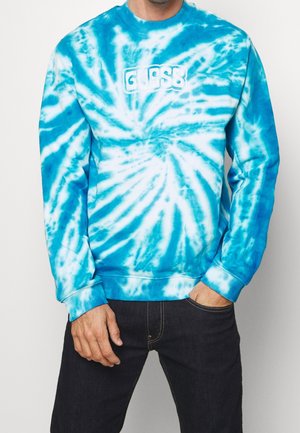 Osoba ubrana w niebiesko-białą bluzę z efektem tie-dye z logo "GUESS" na piersi, w ciemnych dżinsach, z jedną ręką w kieszeni.