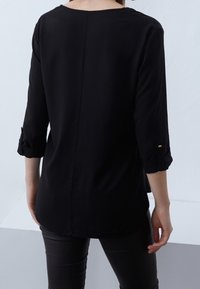 Persona che indossa una maglietta nera a maniche lunghe con le maniche arrotolate e pantaloni neri, in piedi davanti a uno sfondo chiaro e semplice.