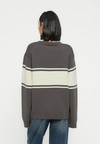 Άτομο με μαύρα μαλλιά μήκους ώμου που φοράει γκρι πουλόβερ με φαρδιές λευκές οριζόντιες ρίγες και μπλε τζιν, γυρισμένο με την πλάτη σε λευκό φόντο.