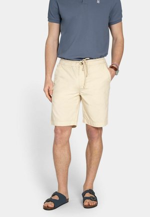 Hombre con pantalones cortos beige con cordón, camiseta polo gris oscuro, sandalias azul marino, reloj y pulseras, de pie con una mano en el bolsillo.