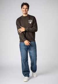 Brown sweatshirt with a graphic print on the left chest, paired with blue jeans and white sneakers. Soft texture, casual fit.