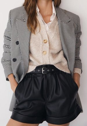Kvinde iført sort- og hvidternet blazer, beige ribstrikket knap kardigan, hvid skjorte og sorte bælte-lædershorts med hænderne i lommerne.