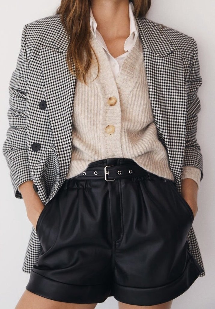 Femme portant un blazer à carreaux noir et blanc, un cardigan beige côtelé boutonné, une chemise blanche et un short en cuir noir ceinturé, mains dans les poches.