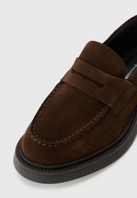 Mocassins en daim marron avec un panneau avant surélevé, des coutures contrastées et un bout arrondi. Semelle en caoutchouc noir avec un bord texturé.