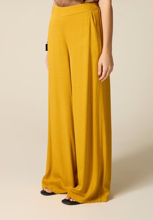 Donna che indossa pantaloni larghi a vita alta color senape con tasche laterali e sandali neri con punta aperta, in piedi su sfondo beige.