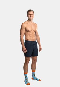 Modell trägt dunkelblaue Shorts und bunte blaue Socken mit Pizza- und Burger-Motiven, steht mit entspannten Armen an den Seiten vor einem grauen Hintergrund.
