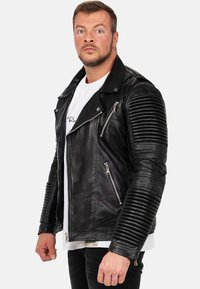 Zwarte leren motorjack met ribbelaccenten op de schouders en mouwen, voorzien van ritssluitingen en zilveren hardware.
