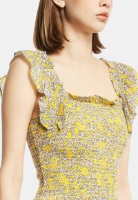 Abito floreale giallo con spalline arricciate, corpetto smock elasticizzato e piccoli motivi floreali bianchi e rosa su tessuto testurizzato.