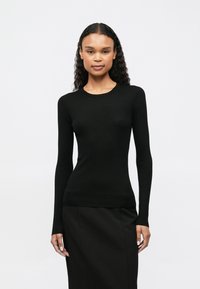 Žena s kovrčavom kosom koja nosi usku crnu majicu dugih rukava od rebraste tkanine i crnu suknju, stoji nasuprot jednostavne svijetle pozadine.