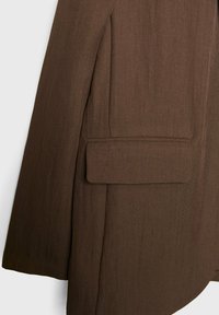 Manche d'un blazer marron sur mesure avec une poche à rabat rectangulaire et des lignes de couture nettes et structurées.