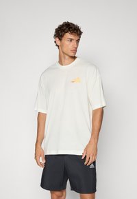 Kremfarget oversized t-skjorte med korte ermer som har en oransje logo, kombinert med svarte shorts som viser en hvit merkevarelogo.