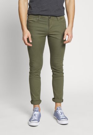 Homme portant un pantalon slim vert olive retroussé aux chevilles avec des baskets en toile bleu clair et une chemise gris foncé.