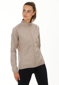 Lätt beige idrottsjacka med hög krage, hel dragkedja och reflekterande detaljer på ärmarna. Slät yta och figursydd design.