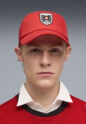 Joven con gorra roja con el emblema de fútbol de Austria y camiseta roja sobre una camisa blanca con cuello, con fondo gris.