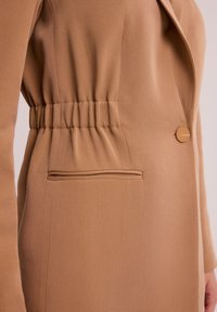 Brązowy blazer z marszczonym talią, jednopunktowym zapięciem i płaską kieszenią. Tkanina ma subtelną teksturę i strukturalny design.